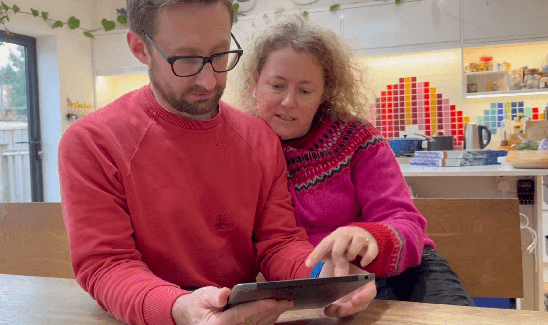 A man in a red sweatshirt using an ipad to answer online safety questions and a woman in a pink sweater pointing at the screen. 