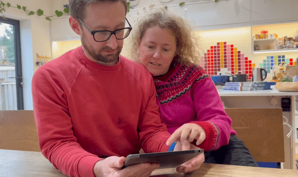 A man in a red sweatshirt using an ipad to answer online safety questions and a woman in a pink sweater pointing at the screen.