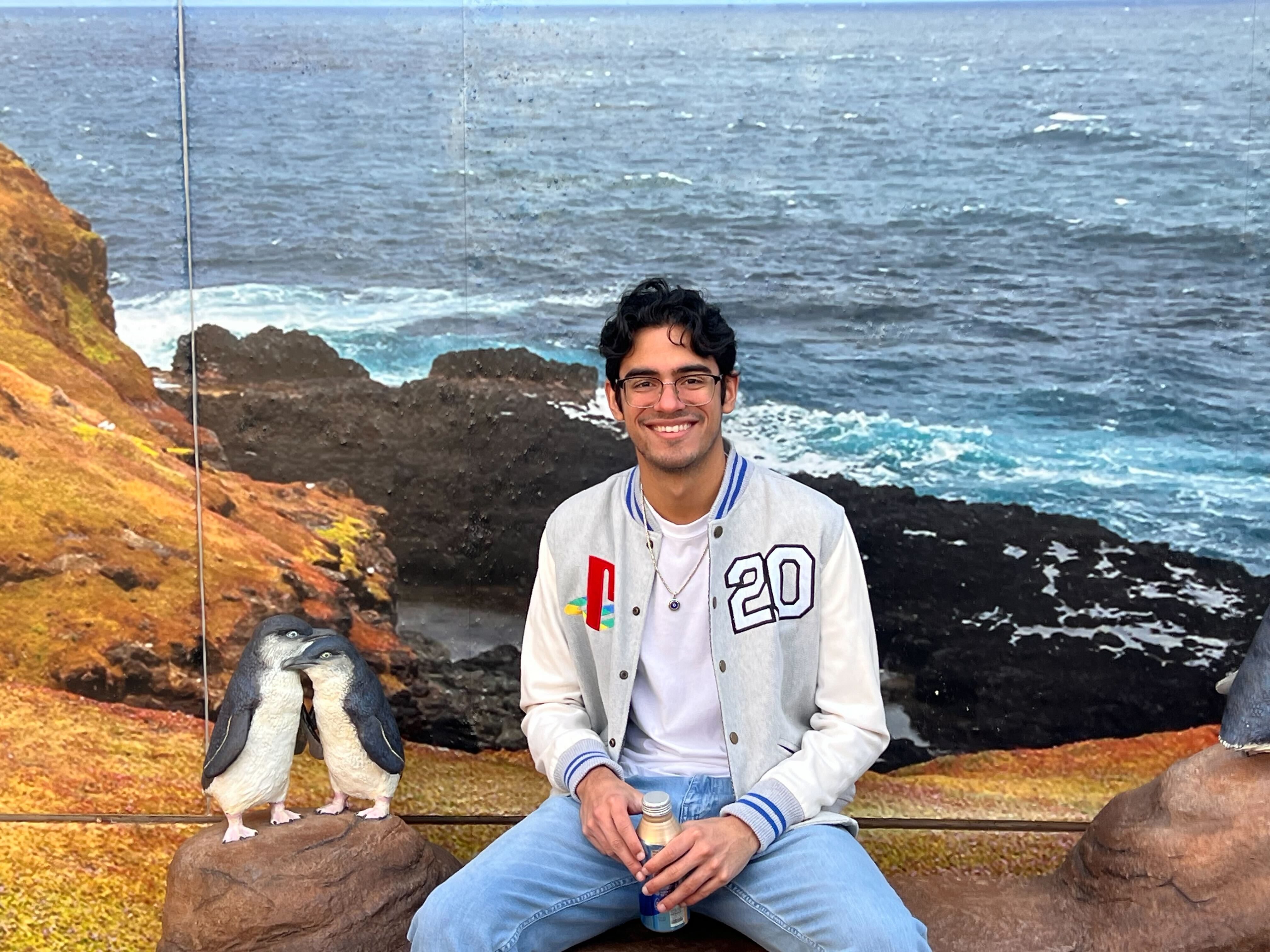 A man with short dark hair, glasses, and a gray PlayStation jacket, sitting in front of a backdrop of an ocean and cliff.
