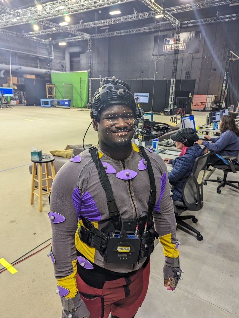 A young man wearing motion capture gear, standing in a large concrete studio.