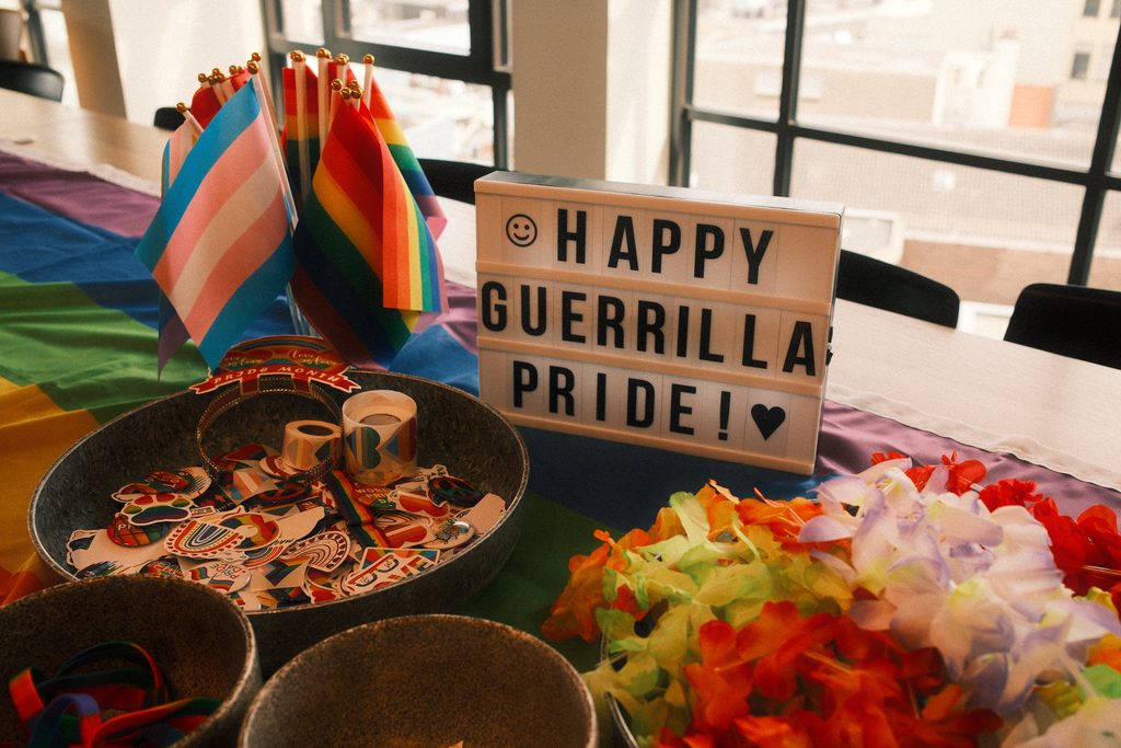 A table with Pride flags and decorations. A sign reads Happy Guerilla Pride!