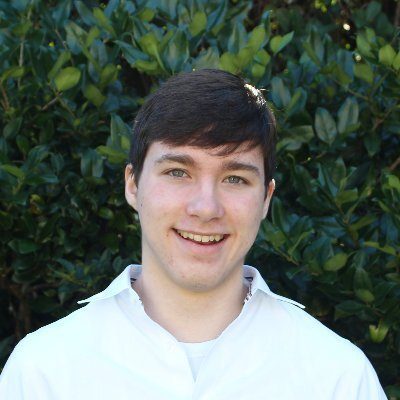 ダークブラウンの短髪に白いシャツを着た若い男性が、緑の葉を背景に微笑んでいる。