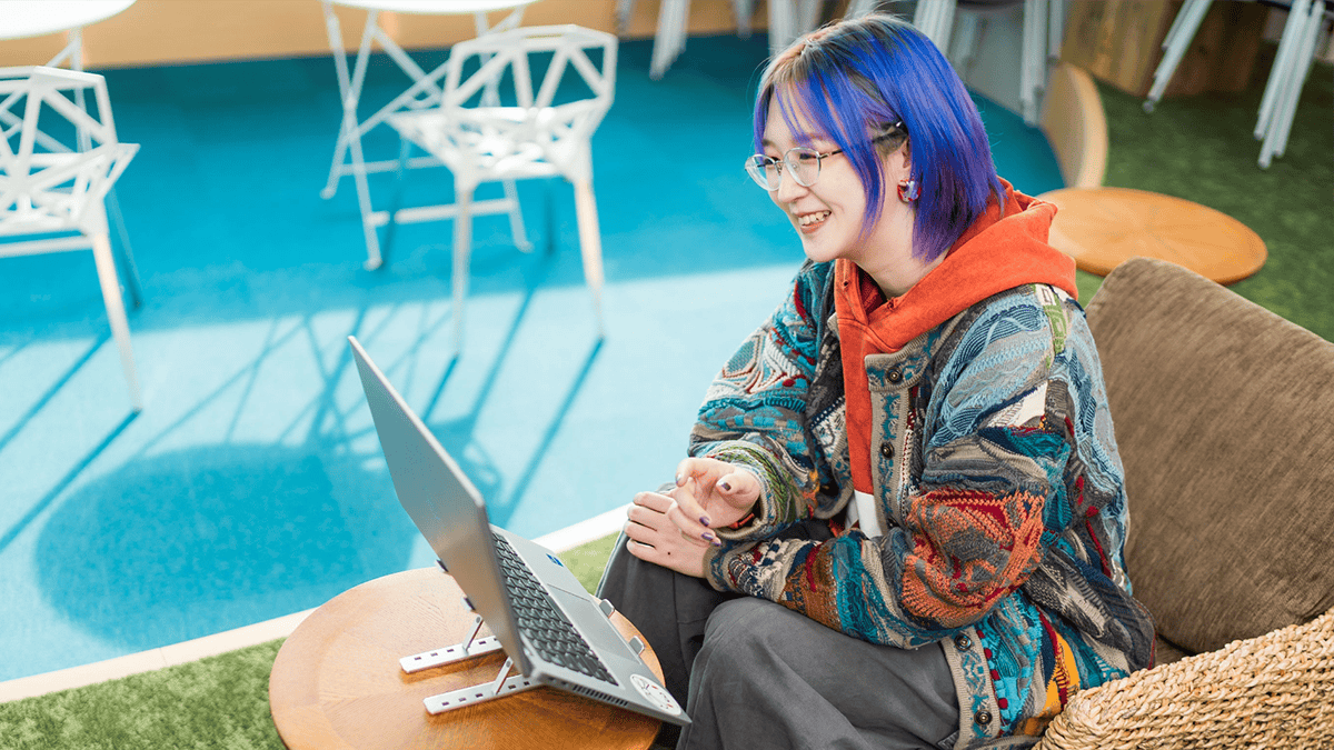 鮮やかな青い髪にカラフルな服を着た女性社員が、茶色のソファに座り、ノートパソコンで会議に参加している。