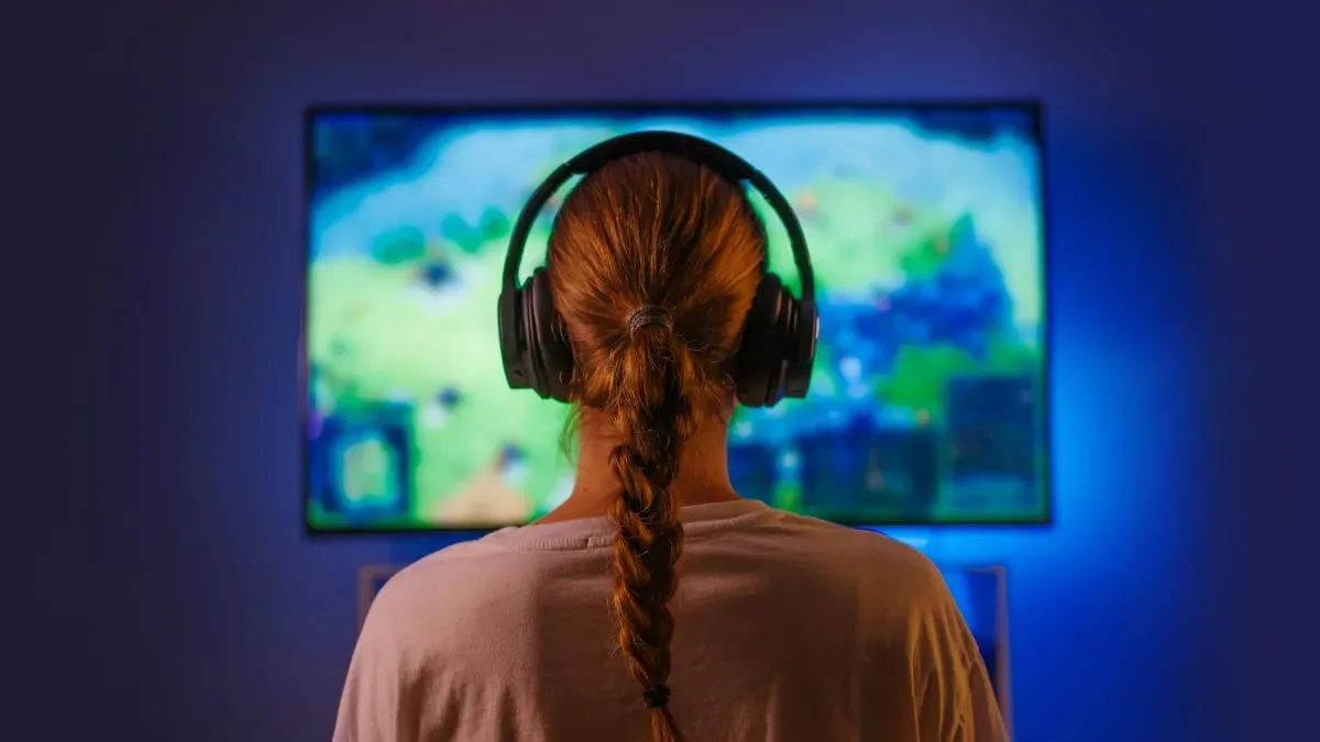 暗い青色の部屋で、大きな黒いヘッドフォンを着用し、テレビ画面を眺めている、長い三つ編みの人物の後ろ姿。